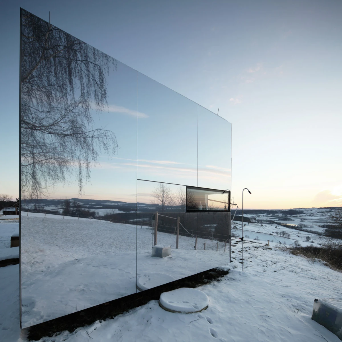 Reflective glass wall of a modern structure in a snow-covered landscape, mirroring bare trees and distant hills at sunset.