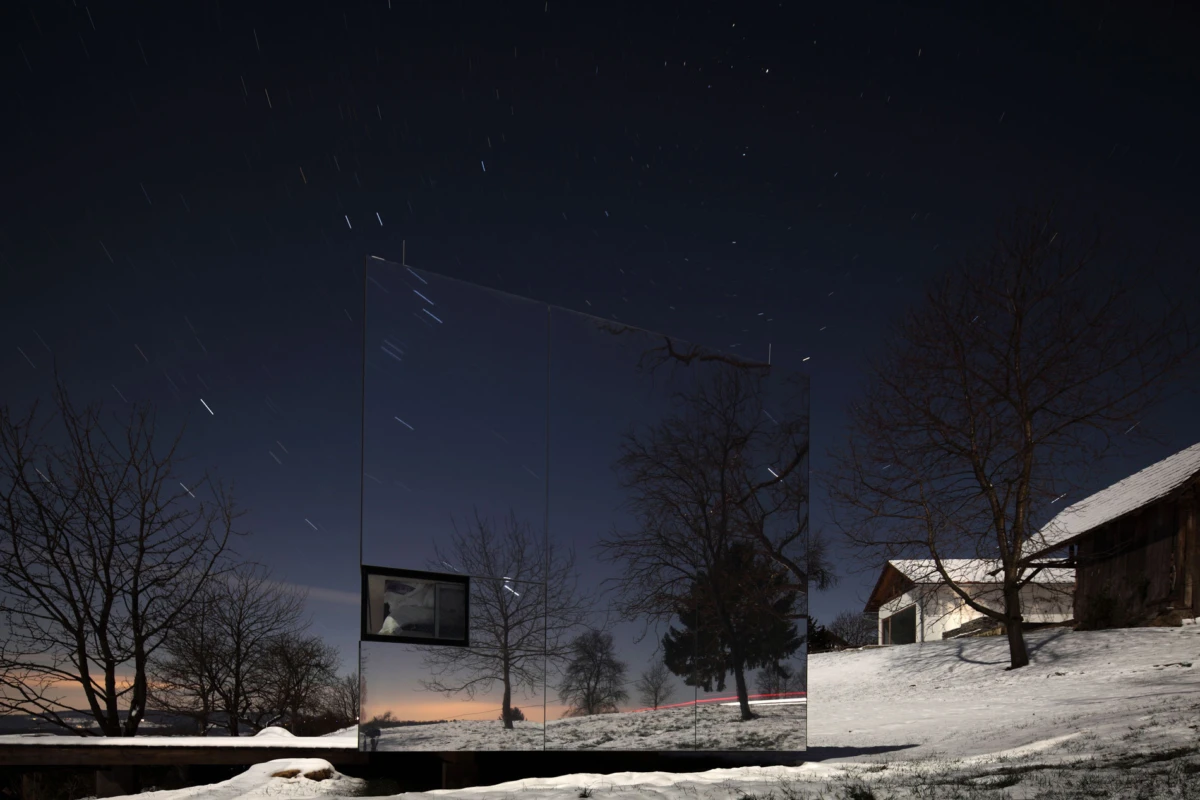 Reflective rectangular panel sculpture in a snow-covered field at night, mirroring bare trees and a barn; star trails in the sky.