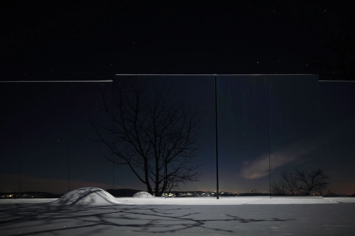 Night snow field with tall reflective panels forming a fence; bare trees reflected on the panels; distant city lights on the horizon.