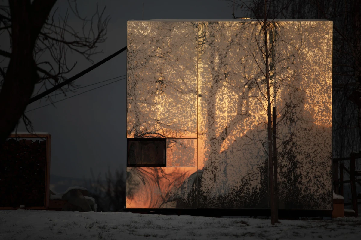Large reflective cube sculpture at dusk, mirroring trees and warm orange light; snowy ground.
