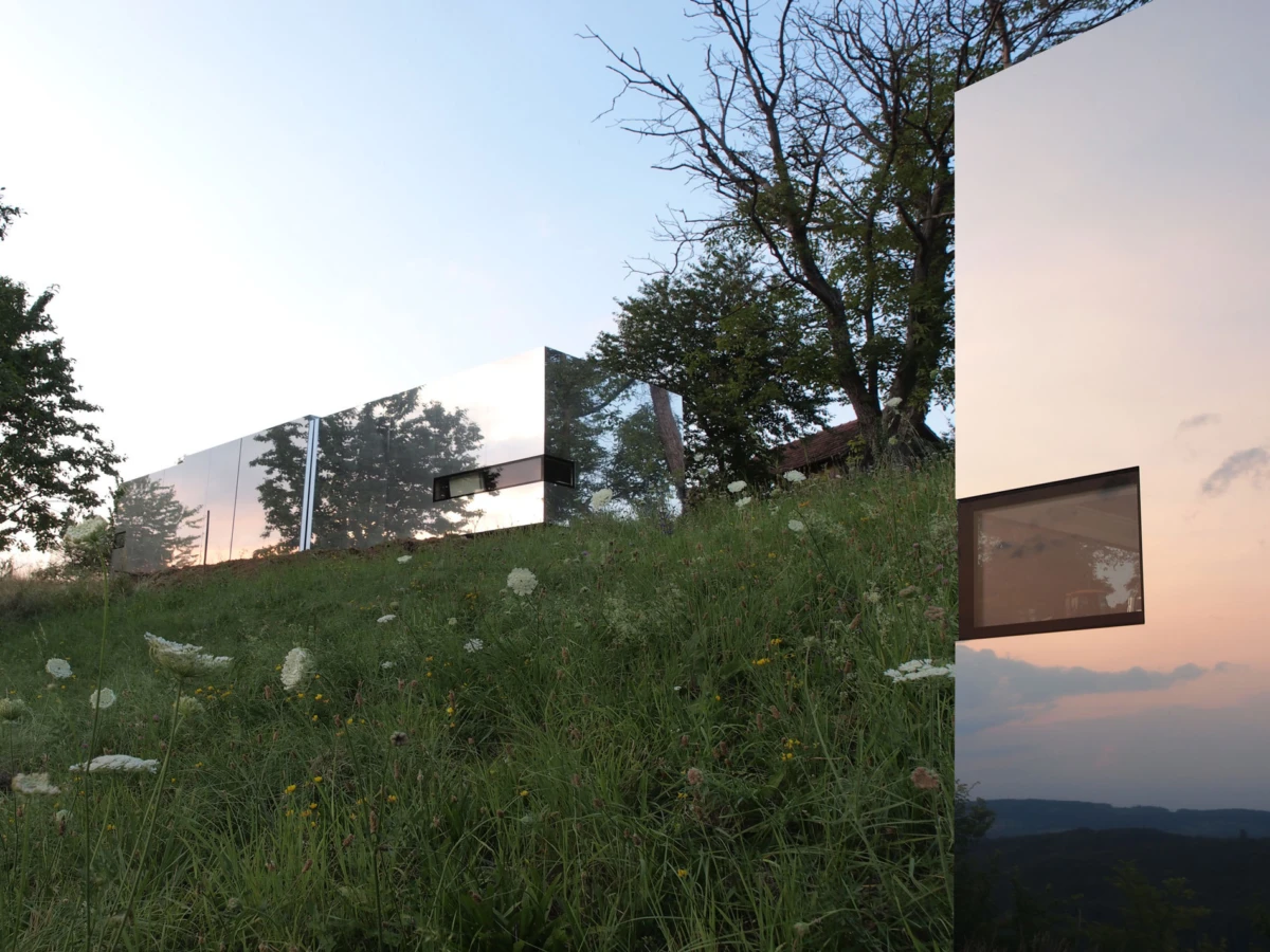 Mirror-clad modern house on a grassy hillside, reflecting trees at sunset.