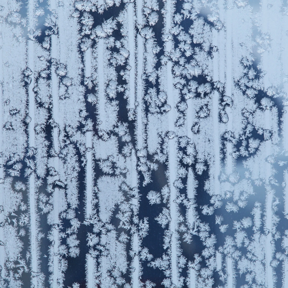 Frost crystals on a window pane forming vertical, fern-like patterns.