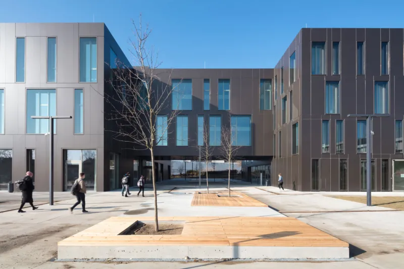 Modern office complex with a central pedestrian plaza, leafless trees in wooden planter boxes, and people walking.