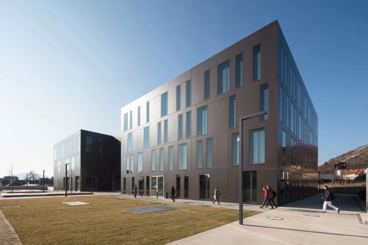 Four-story modern office building with glass windows; pedestrians walk toward the entrance on a sunny day.