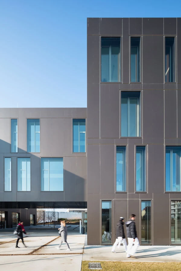 Modern dark-gray office building with tall rectangular windows; pedestrians walk along a sunny sidewalk beneath a blue sky.