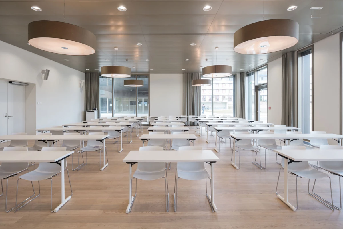Empty modern classroom with rows of white desks and plastic chairs, large circular pendant lights, and floor-to-ceiling windows.