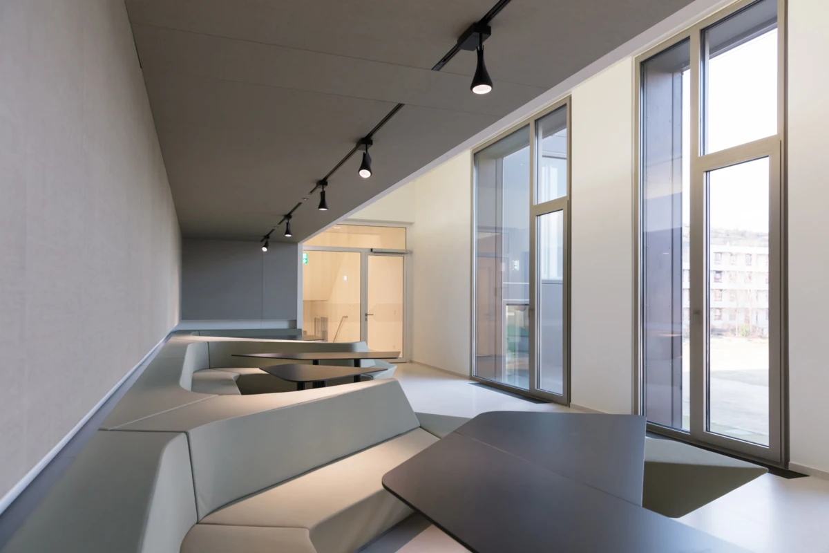 Modern interior lounge with pale gray modular seating and dark rectangular tables along a wall of tall windows; track lighting overhead.