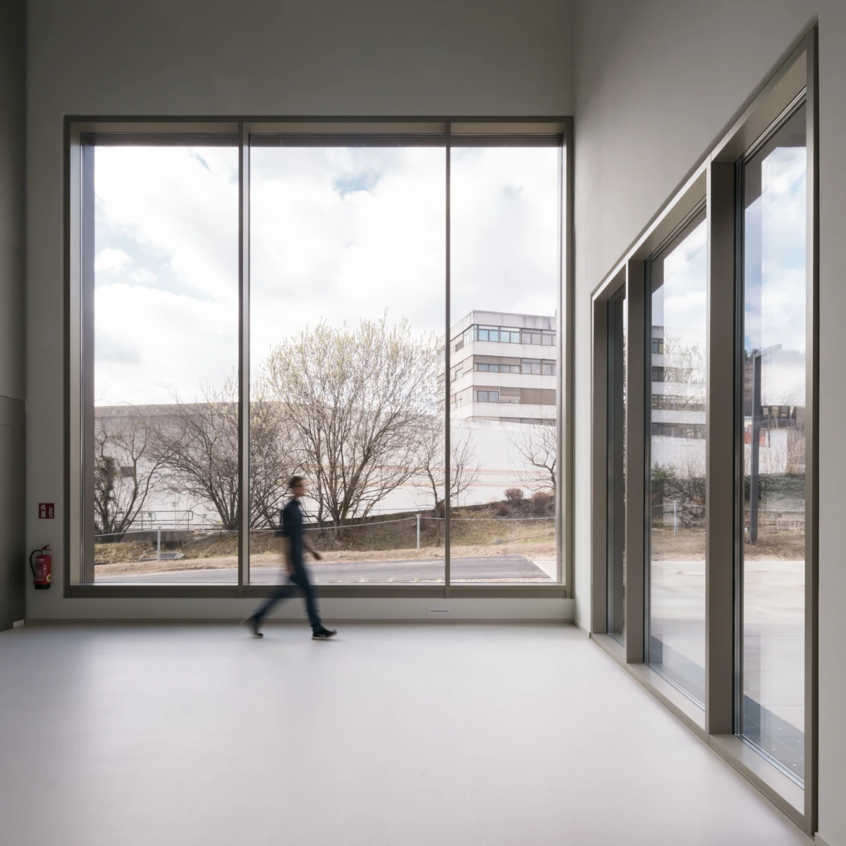 Blurred person walking across a bright, empty modern room with floor-to-ceiling glass walls; outside, leafless trees and a building.