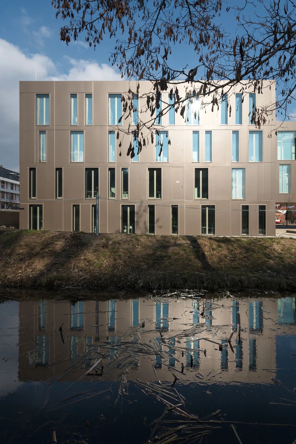 Beige modern office building stands beside a calm canal; its reflection in the water is framed by overhanging bare branches.