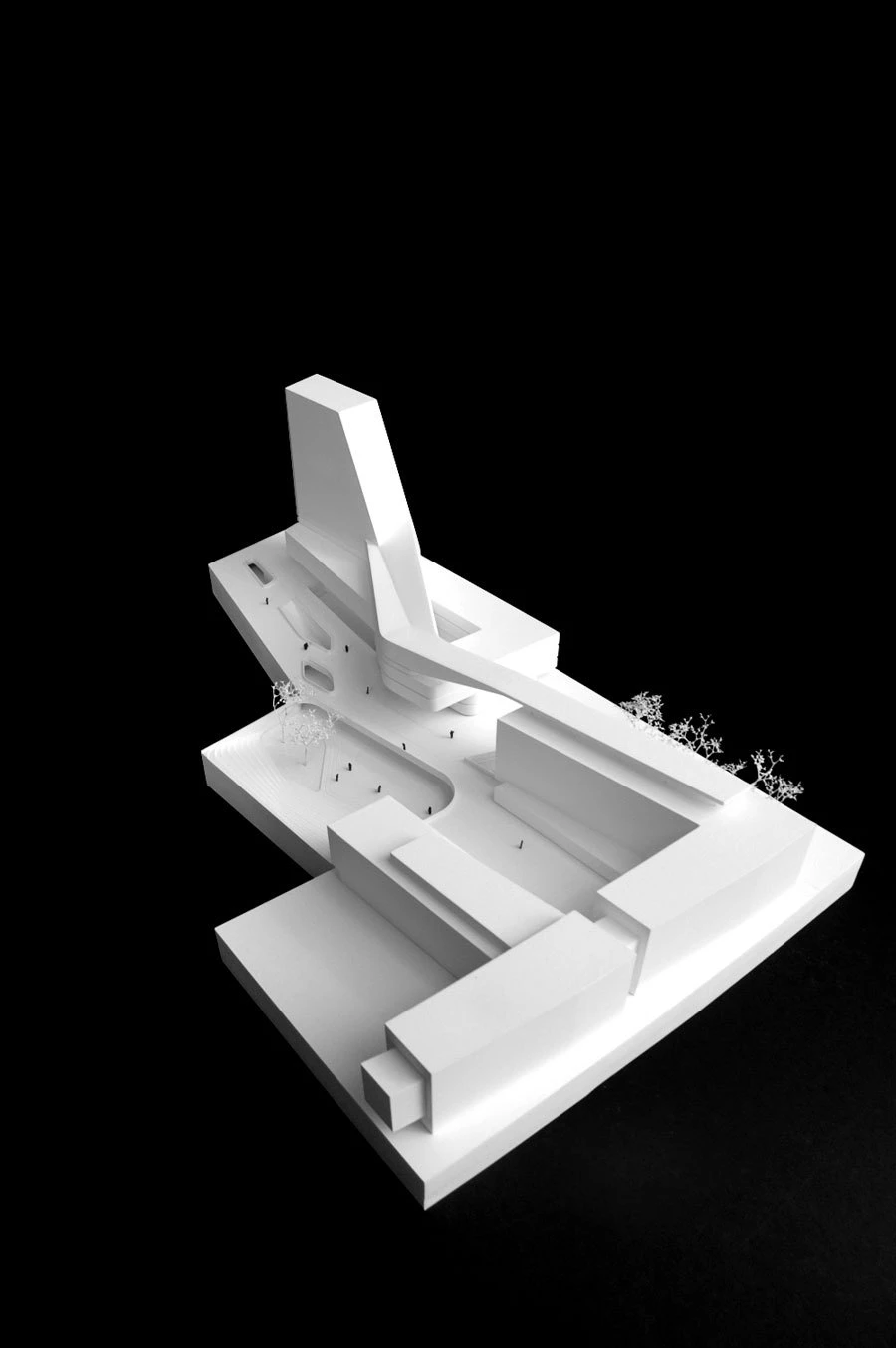 White architectural model of a modern complex featuring a tall tower, angled ramp, stepped platforms, and trees in a plaza against a black background.