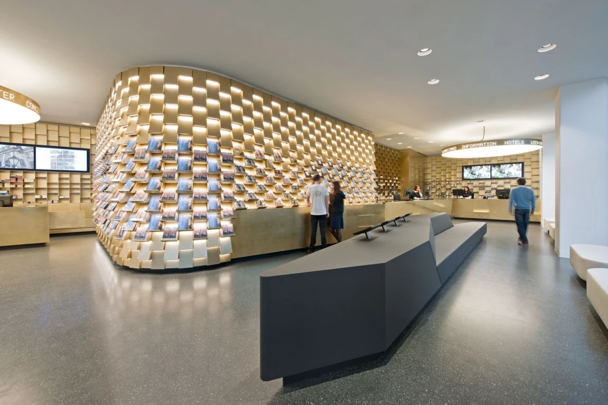 Curved wooden brochure wall at a modern information center; visitors browse brochures near a reception desk.
