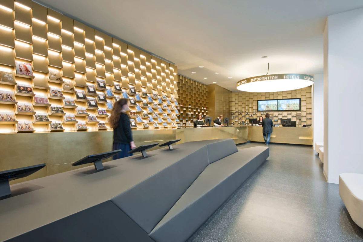 Hotel lobby information desk; staff assist guests, magazine wall along wooden shelves, gray modular seating in foreground.