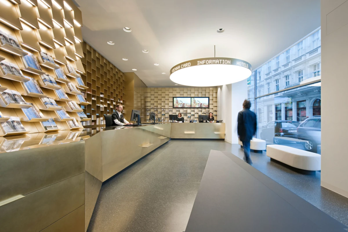Information desk in a modern hotel lobby; staff at computers, magazine wall to the left, and a person walking past a large window.