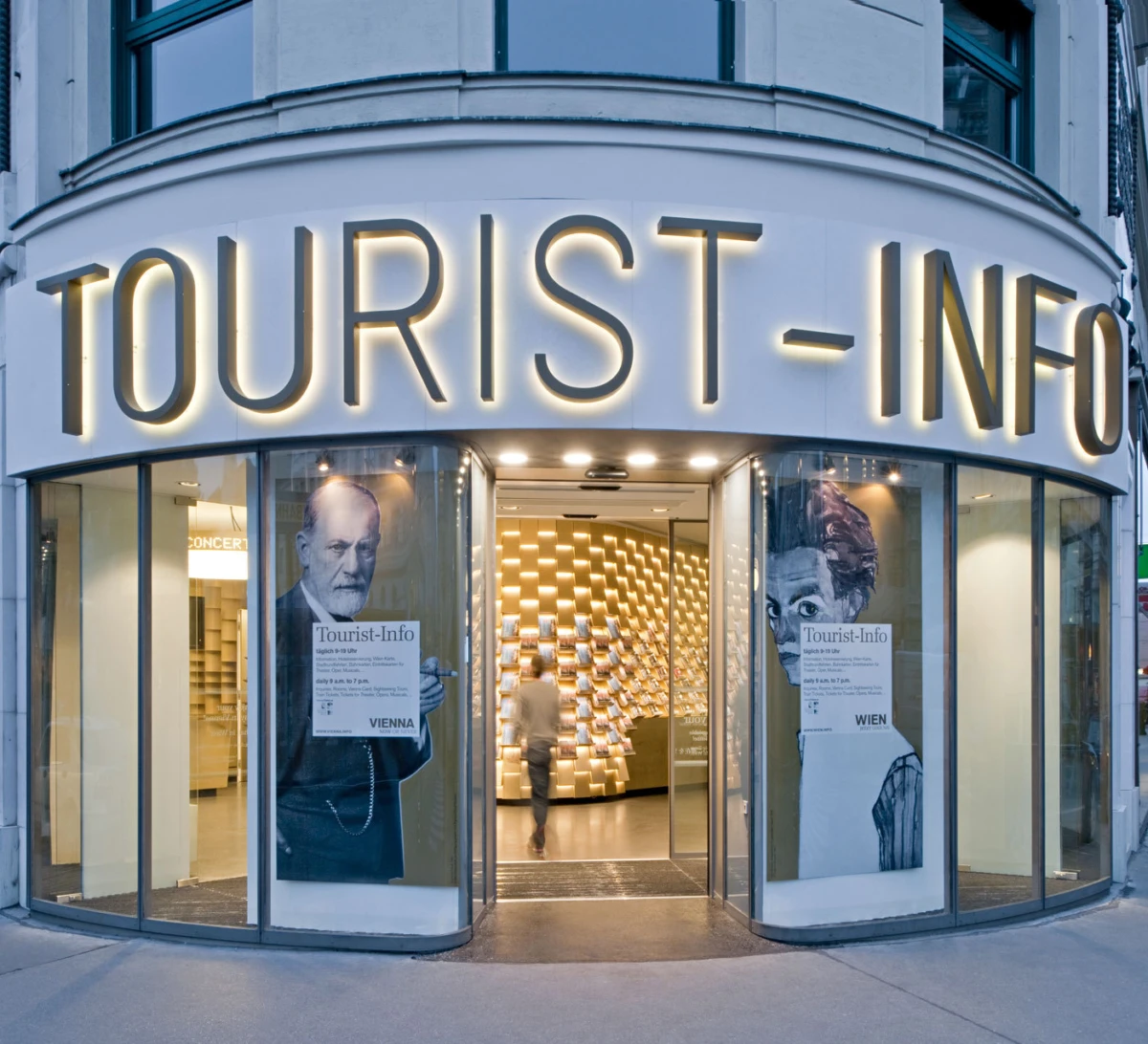 Front of a tourist-information center with a large illuminated 'TOURIST-INFO' sign; a person enters through glass doors, flanked by portraits.