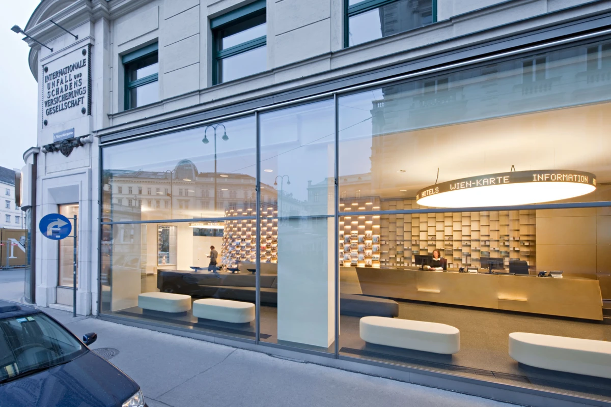 Glass-front hotel reception desk in a modern lobby; receptionist behind the counter, white ottomans, and glass storefront facing a city street.