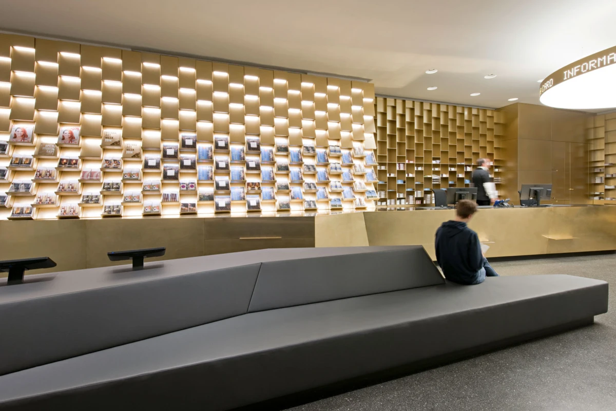 Person seated on a gray modular bench in front of a modern information desk with wall-mounted magazine racks.