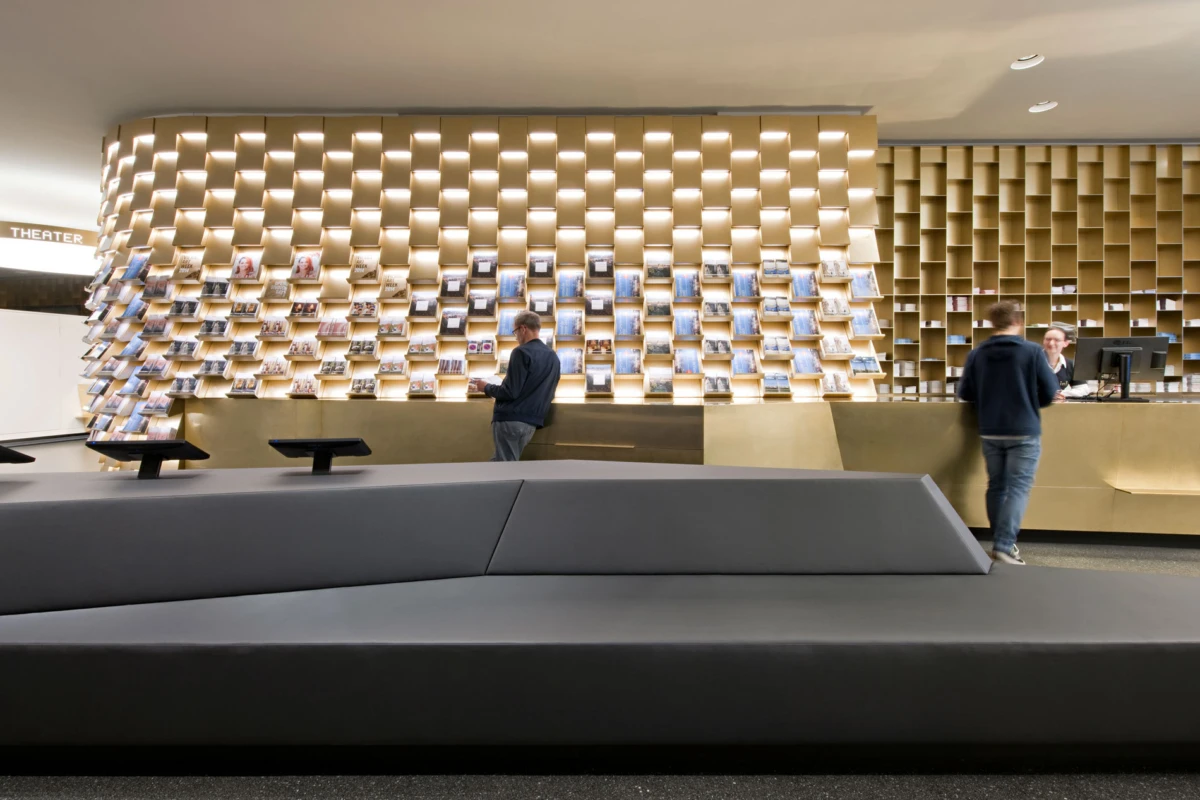 Person in a dark jacket browses magazines on a curved magazine wall inside a modern theater lobby near a beige reception desk.