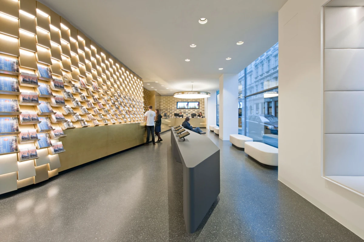 Two people browse magazines at a beige counter in a modern lobby with a magazine display wall and window seating.