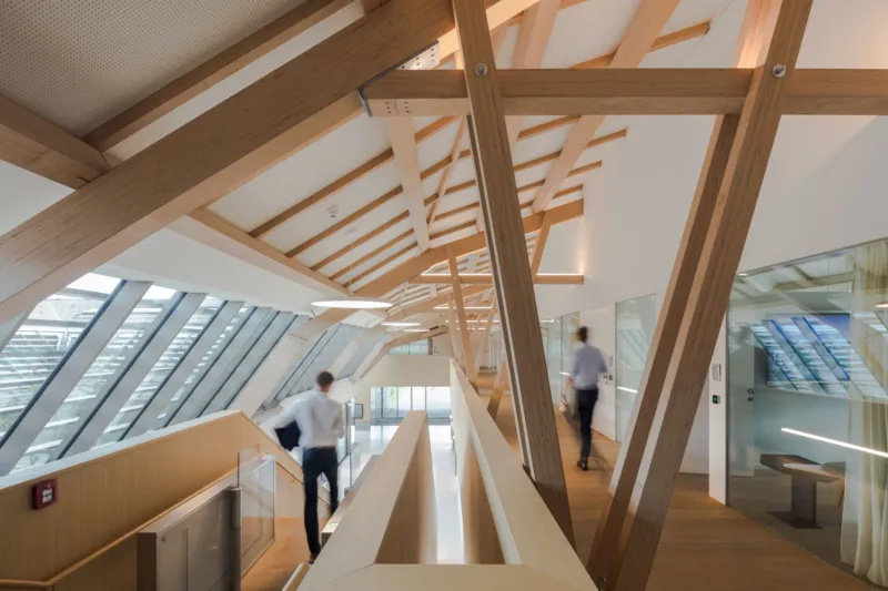 Two professionals walk through a modern atrium featuring exposed timber beams, wooden floors, and slanted glass walls.