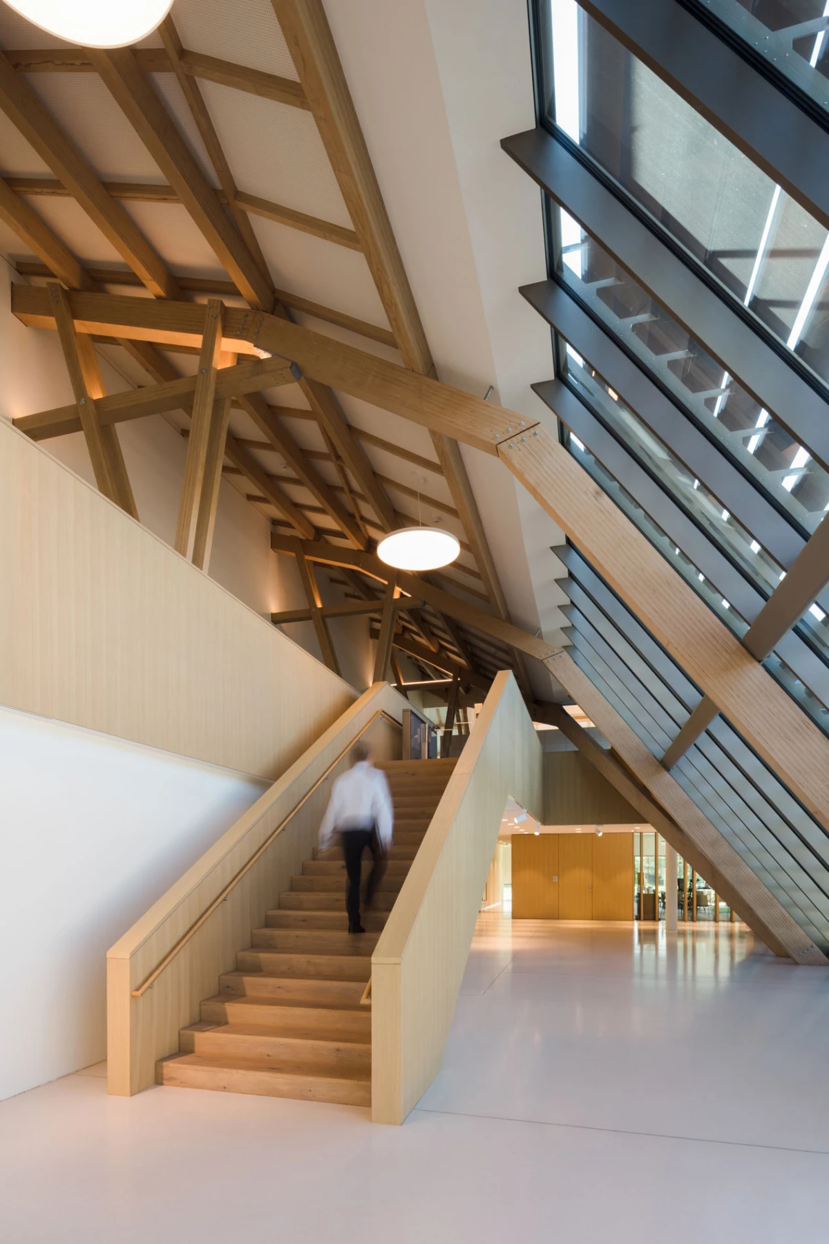 Person ascending a wide wooden staircase in a modern interior with exposed timber beams and a glass wall.