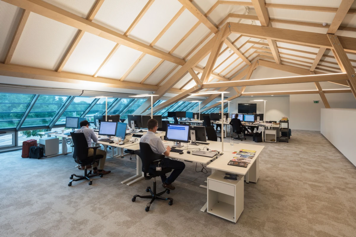 Open-plan attic office with exposed timber beams; employees at desks using desktop computers near large glass windows.