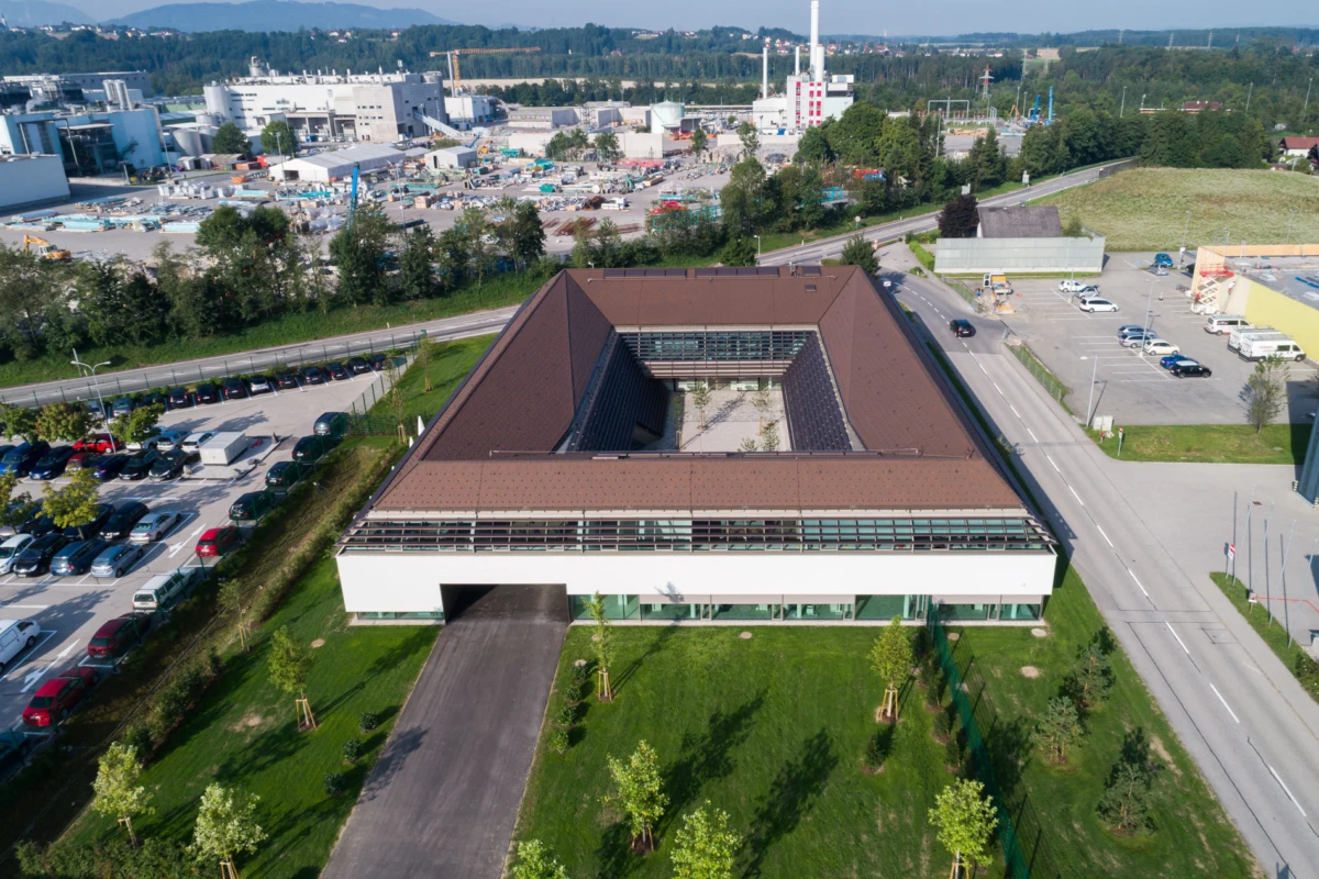 Aerial view of a modern U-shaped office building with a central courtyard and brown roof, surrounded by parking lots, roads, and green lawns.