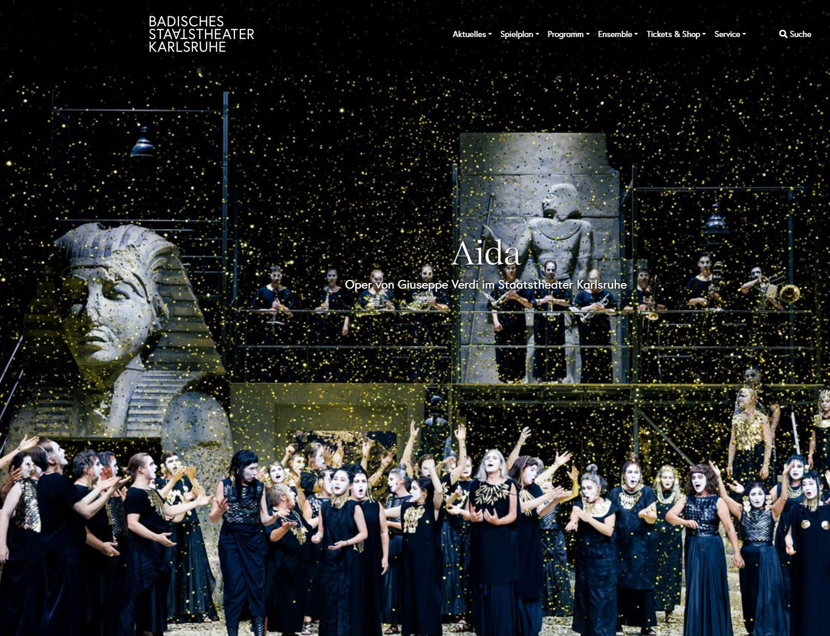 Chorus and orchestra perform Aida on stage at Badisches Staatstheater Karlsruhe, with Egyptian statues and gold confetti.