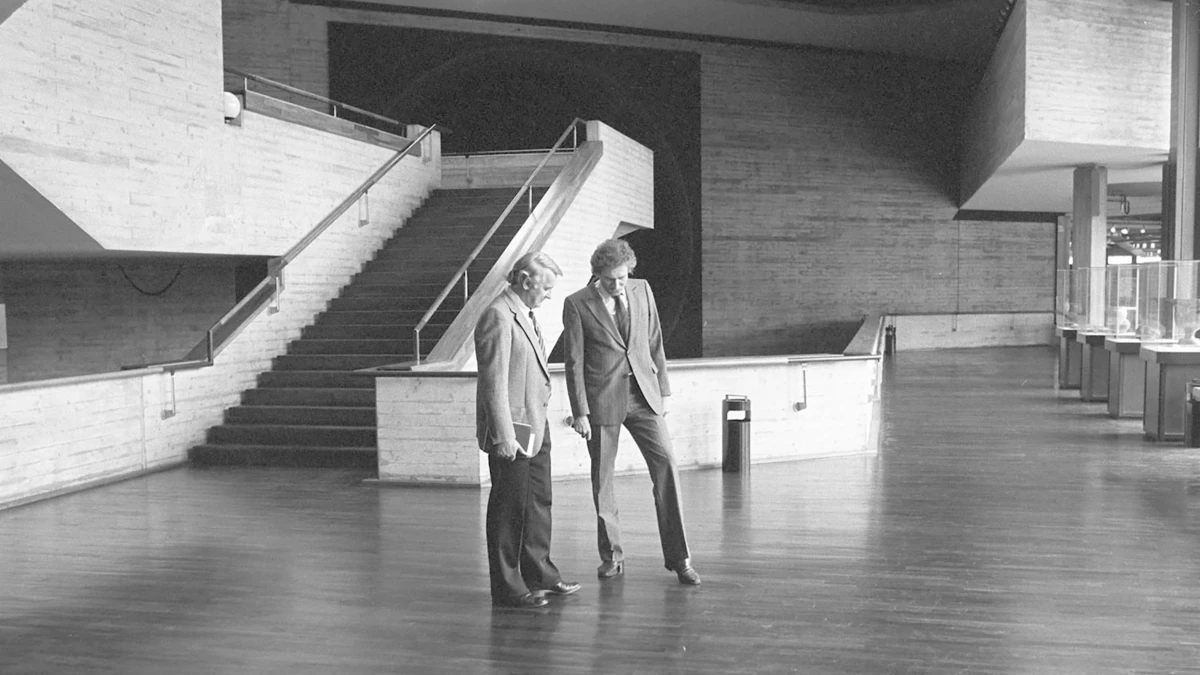 Two men in business suits stand on a polished wooden floor in a spacious modern atrium beside a staircase, looking down.