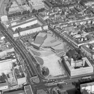 Aerial view of a polygonal-roofed building complex in an urban block, with cars circulating around a circular roundabout and surrounding streets.