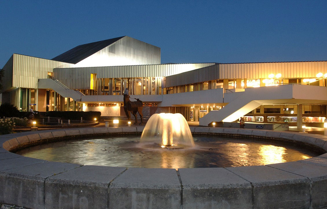 Illuminated fountain spouting water in circular pool in front of a modern, multi-level building at dusk; horse statue visible in background.