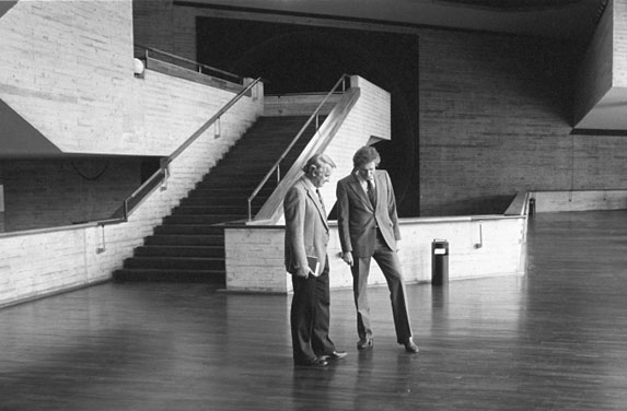 Two people in business attire walk together through a large, empty, modern interior with brick walls and a staircase.