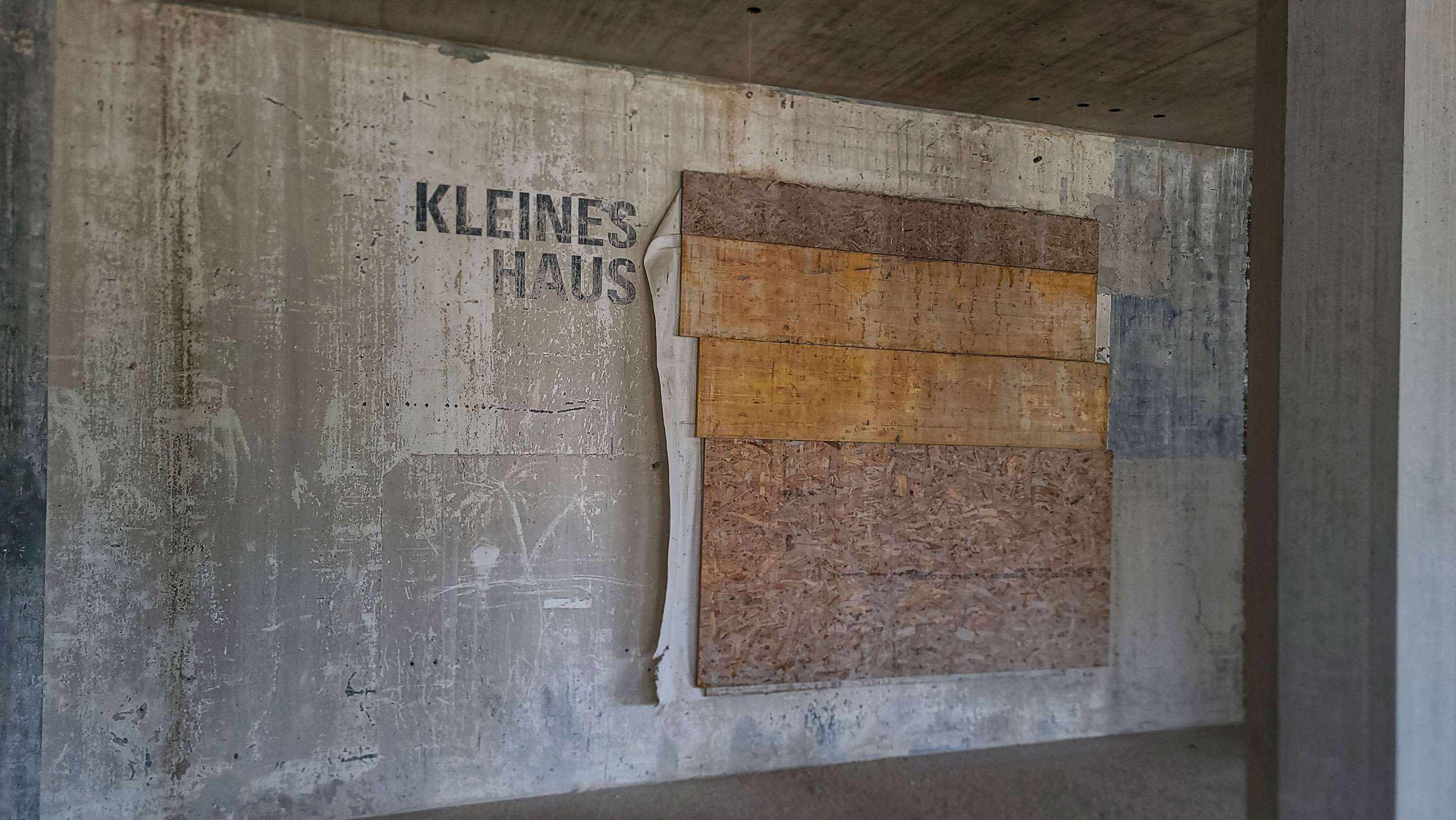 Concrete wall with KLEINES HAUS; stacked plywood panels attached, in an unfinished interior construction space.