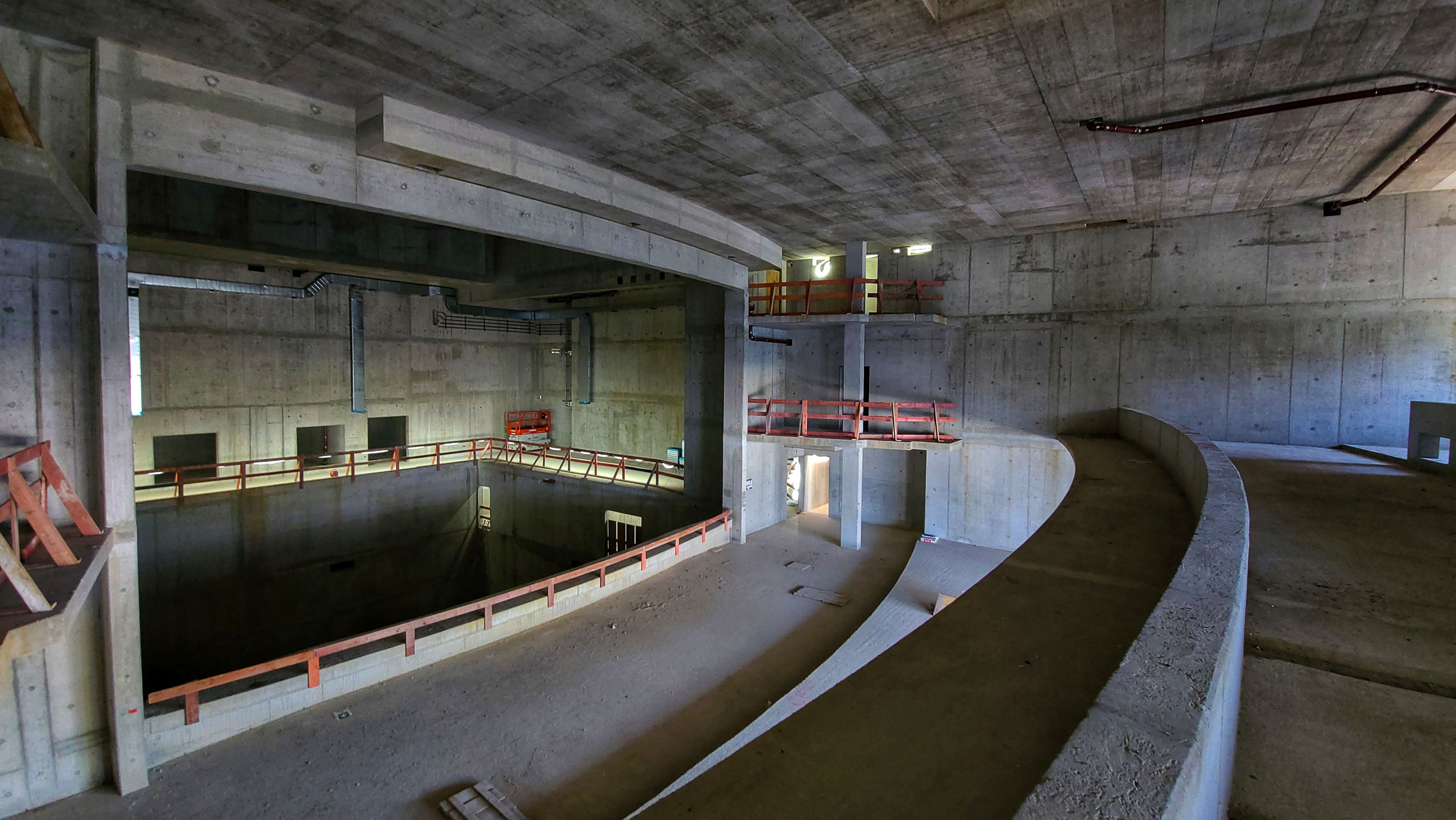 Partially finished concrete construction site with a curved ramp circling a deep open pit, red guard rails, and elevated platforms.