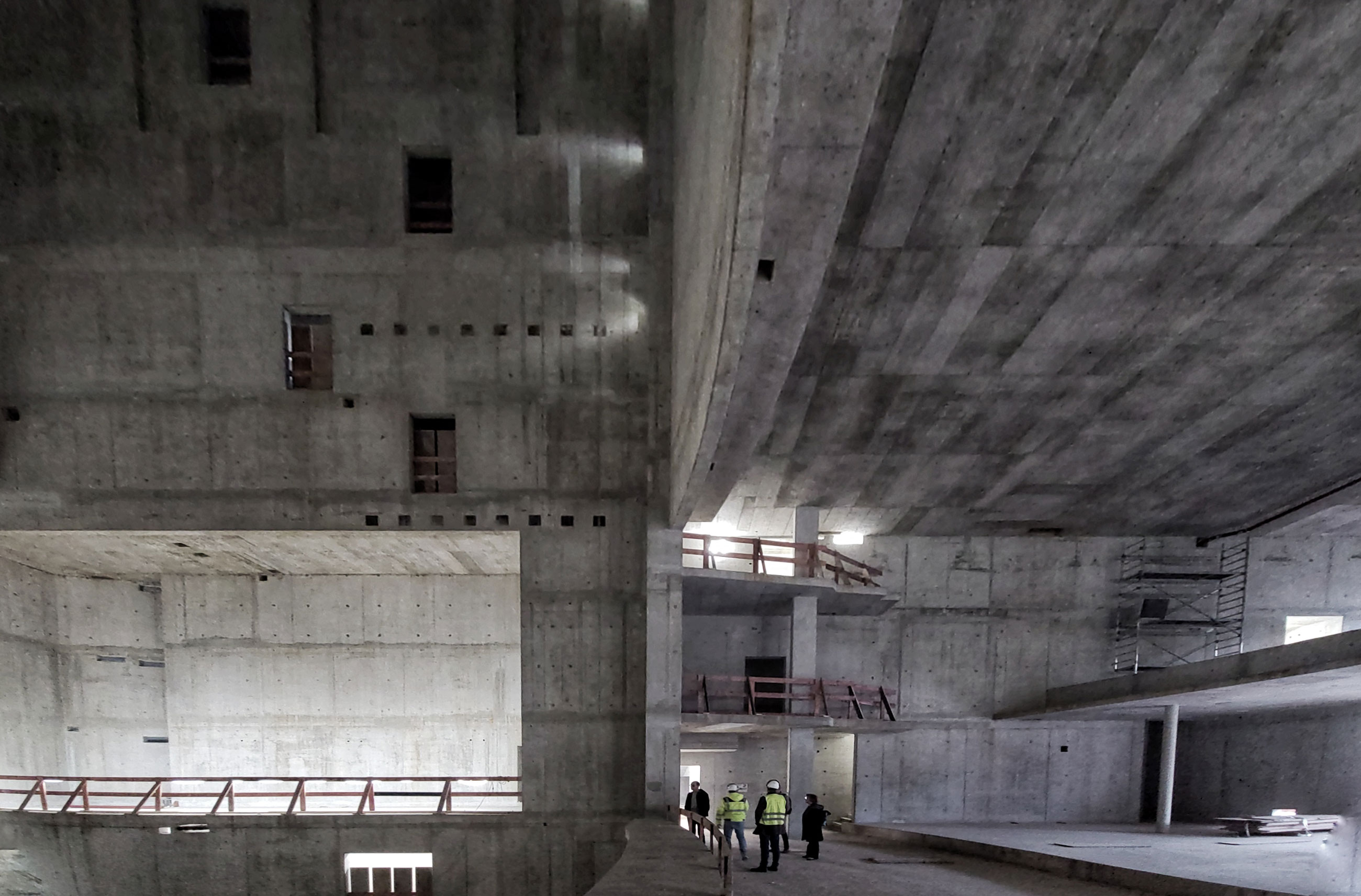 Group of construction workers in yellow safety vests walking inside a vast unfinished concrete building with multiple levels and ramps.