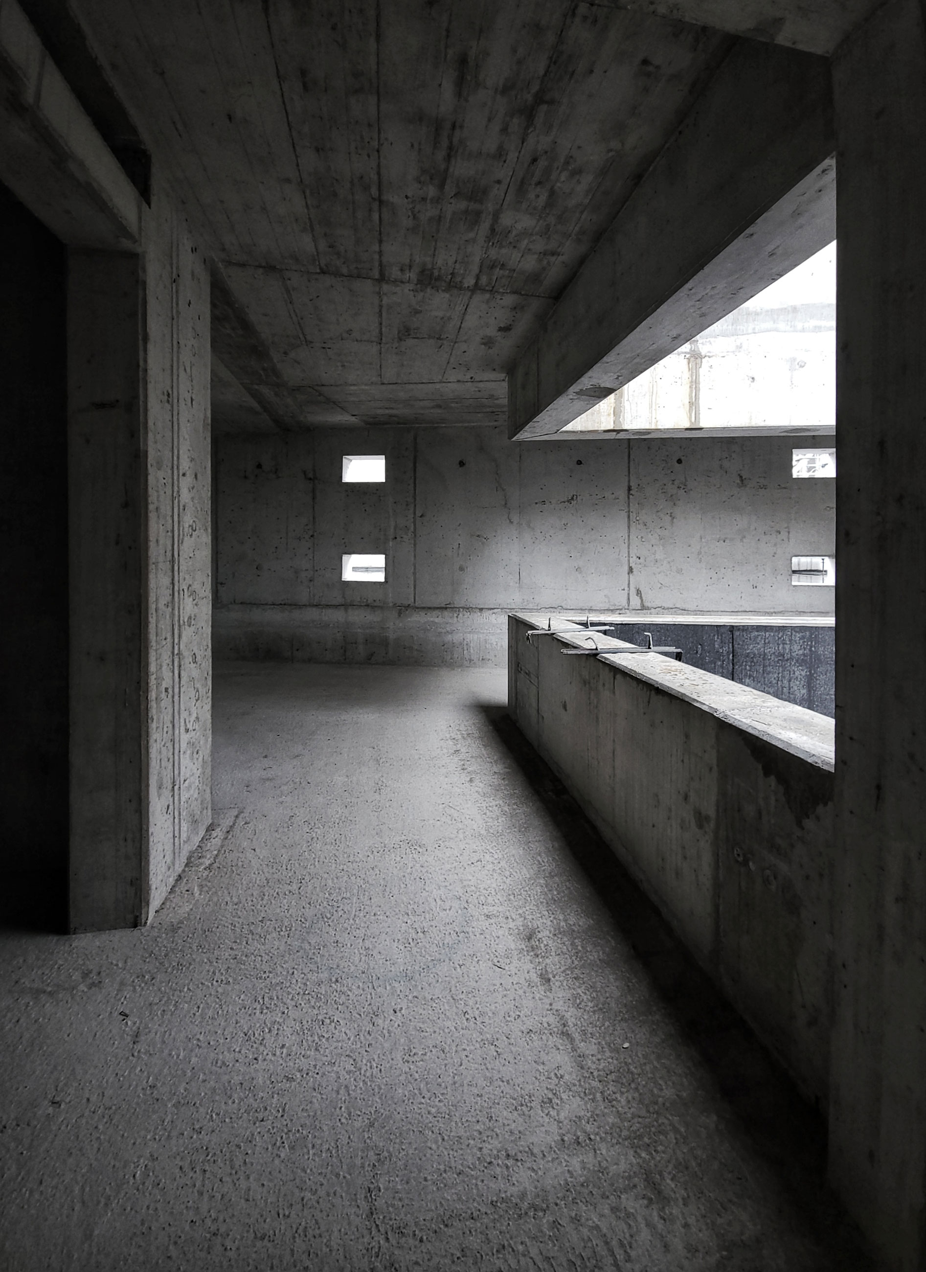 Empty brutalist concrete interior: long corridor with columns, small rectangular windows, and a concrete railing along a raised ledge.