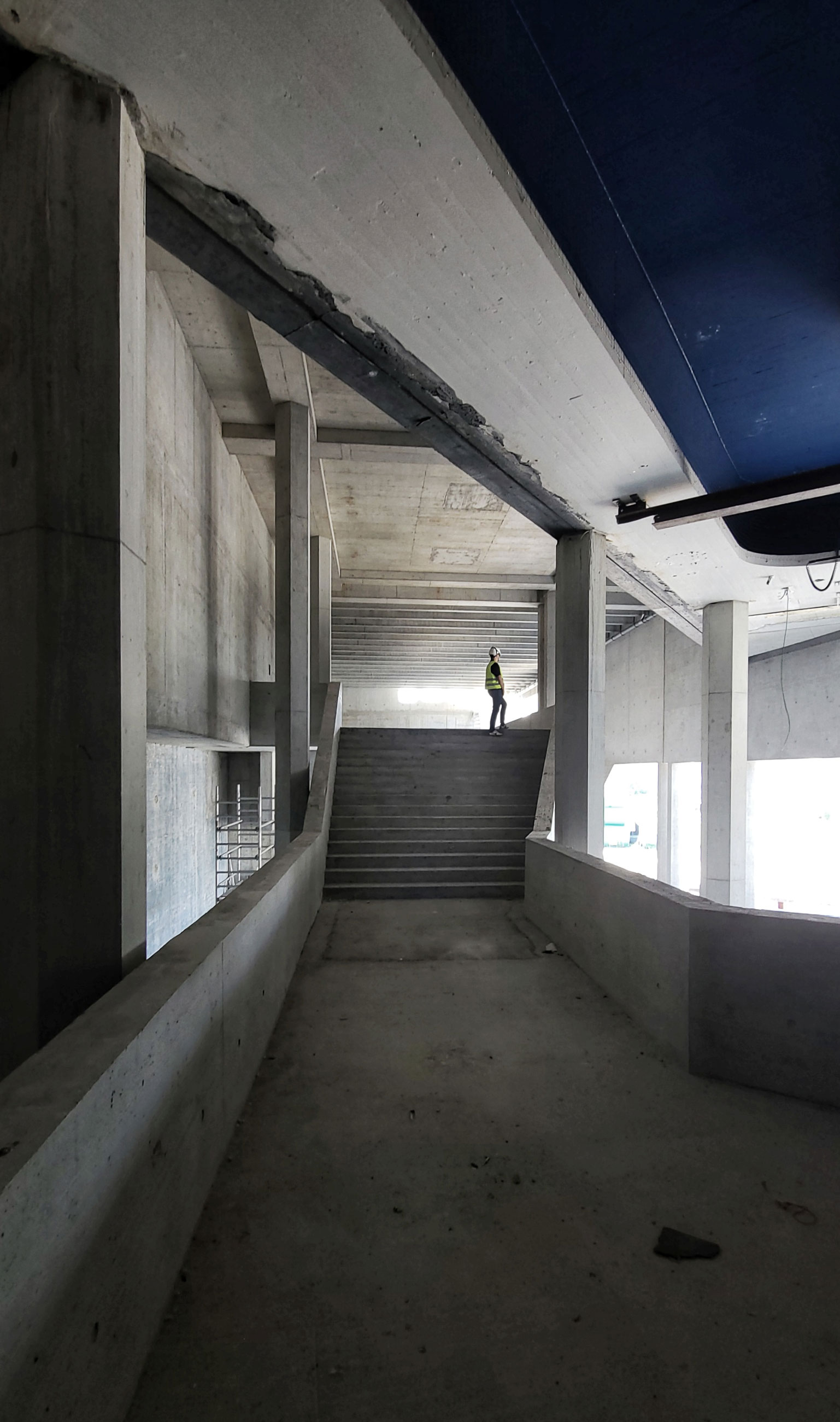 Construction worker in safety vest and hard hat stands on concrete stairs inside unfinished building.