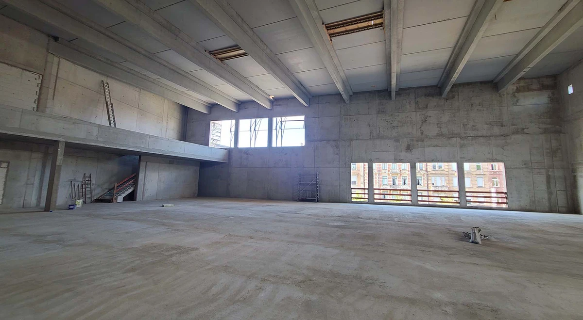 Empty concrete warehouse under construction with a left-side mezzanine, staircase, scaffolding, and large window openings revealing buildings outside.