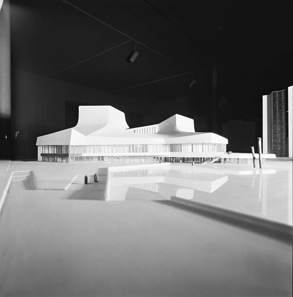 Scale architectural model of a modern white building with sweeping, sculptural roofs on a reflective base in a dark studio.