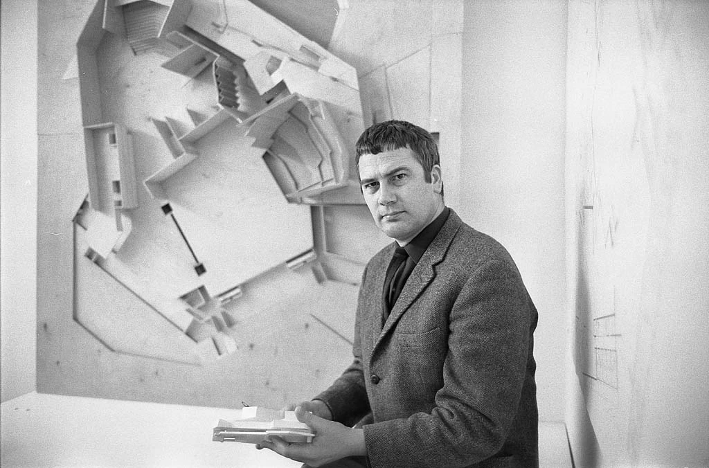 Architect in blazer seated, holding architectural models in a studio with a large geometric wall sculpture.