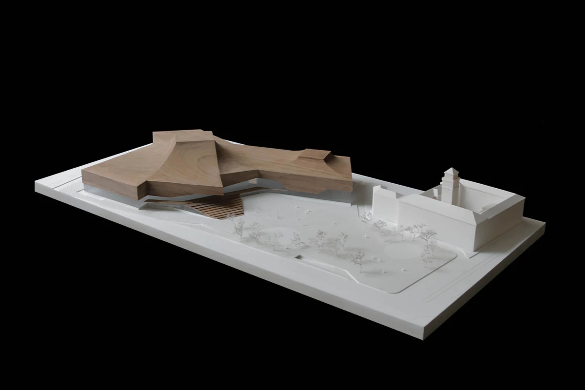 Architectural scale model: irregular wooden mass on a white base, with a separate white block building and a tower on the right.