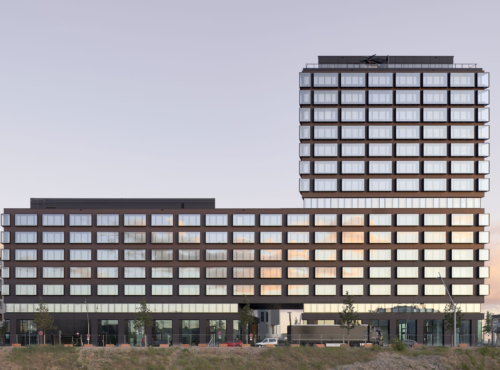 Modern office complex with a low-rise wing connected to a tall glass tower; urban street level with trees and parked cars at sunset.