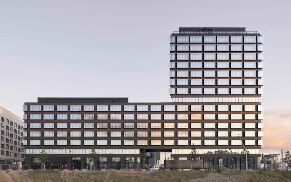 Modern office complex with a low-rise wing connected to a tall glass tower; urban street level with trees and parked cars at sunset.