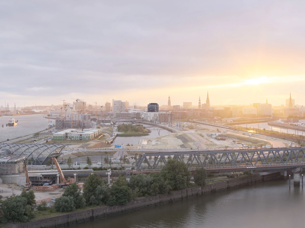 Sunset over a riverfront cityscape with a metal truss bridge crossing the water and a construction site with cranes in the foreground.