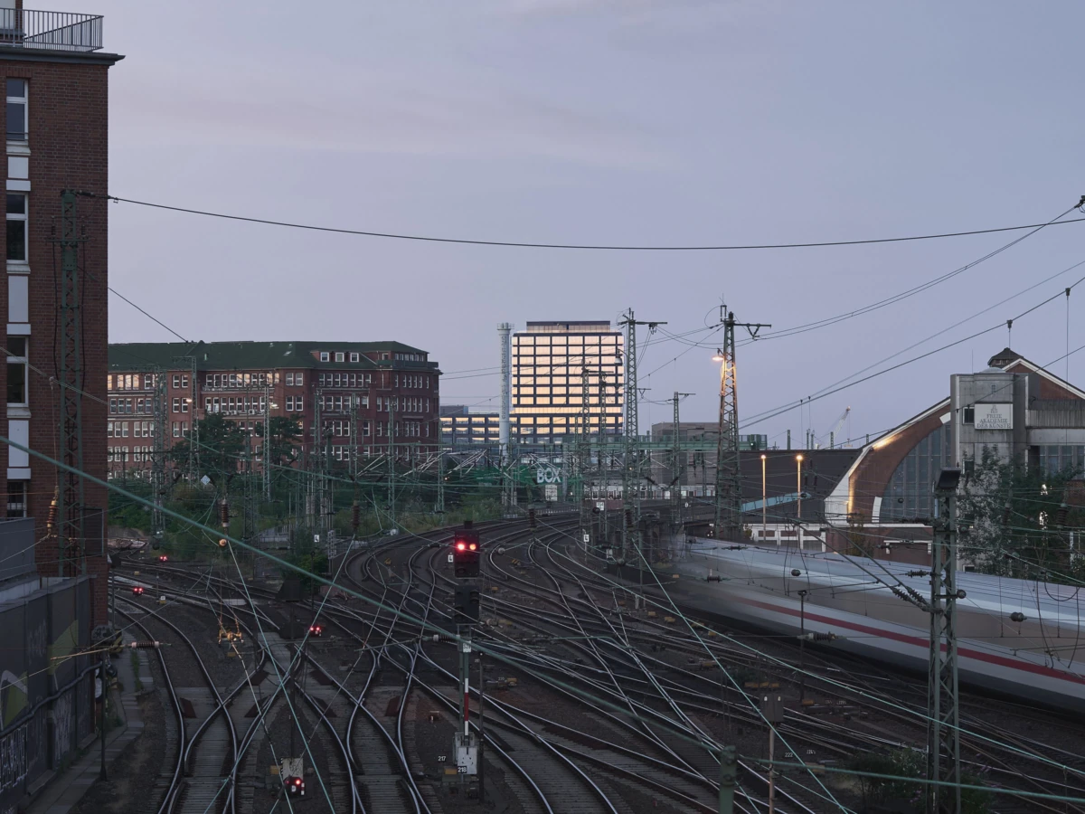 Urban railway yard with tangled curved tracks and overhead lines; a white passenger train on the right and brick buildings in the background.