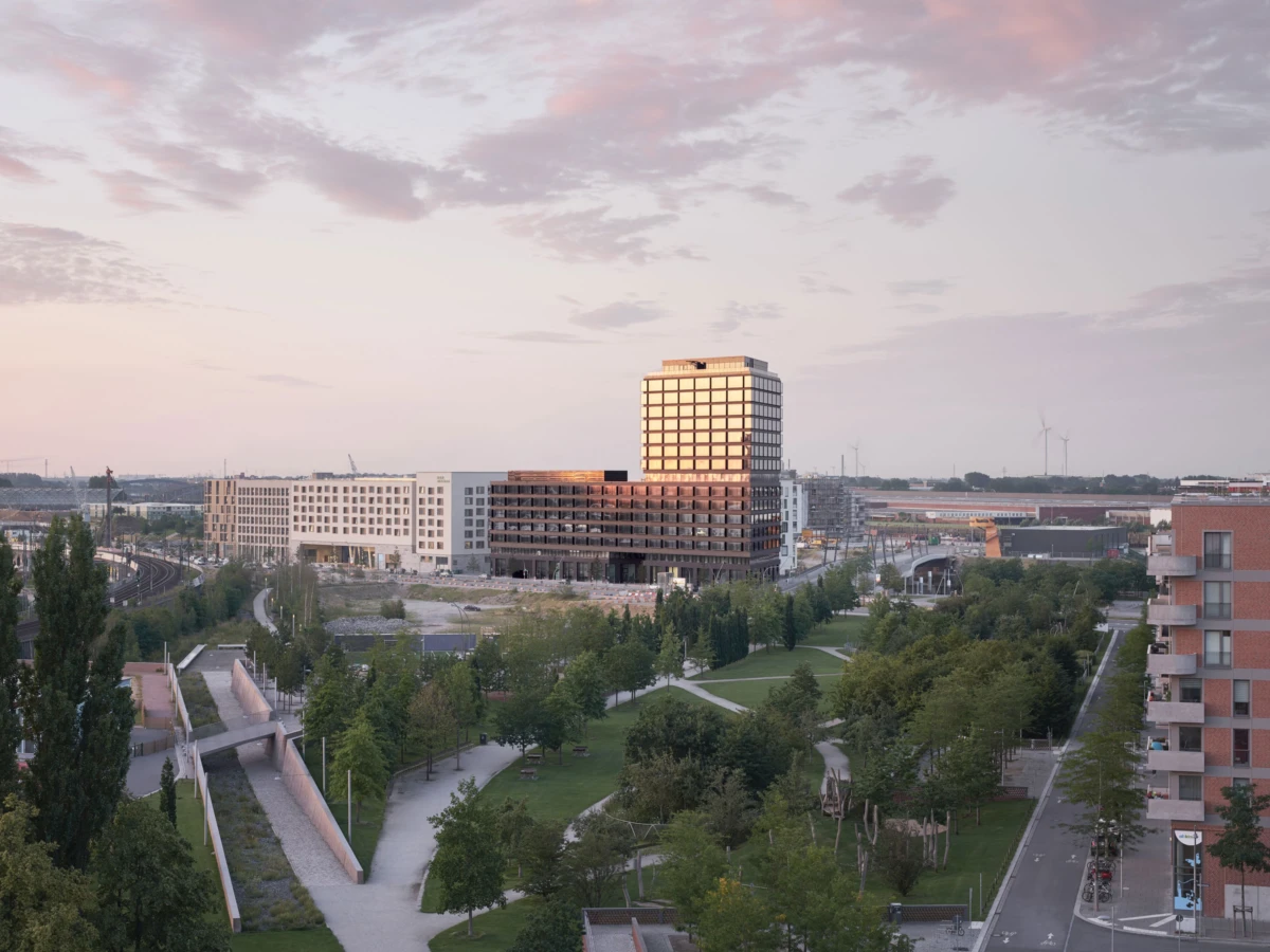 Center-dominant glass-office tower reflecting sunset, set among modern buildings and a green, tree-filled park with winding paths.