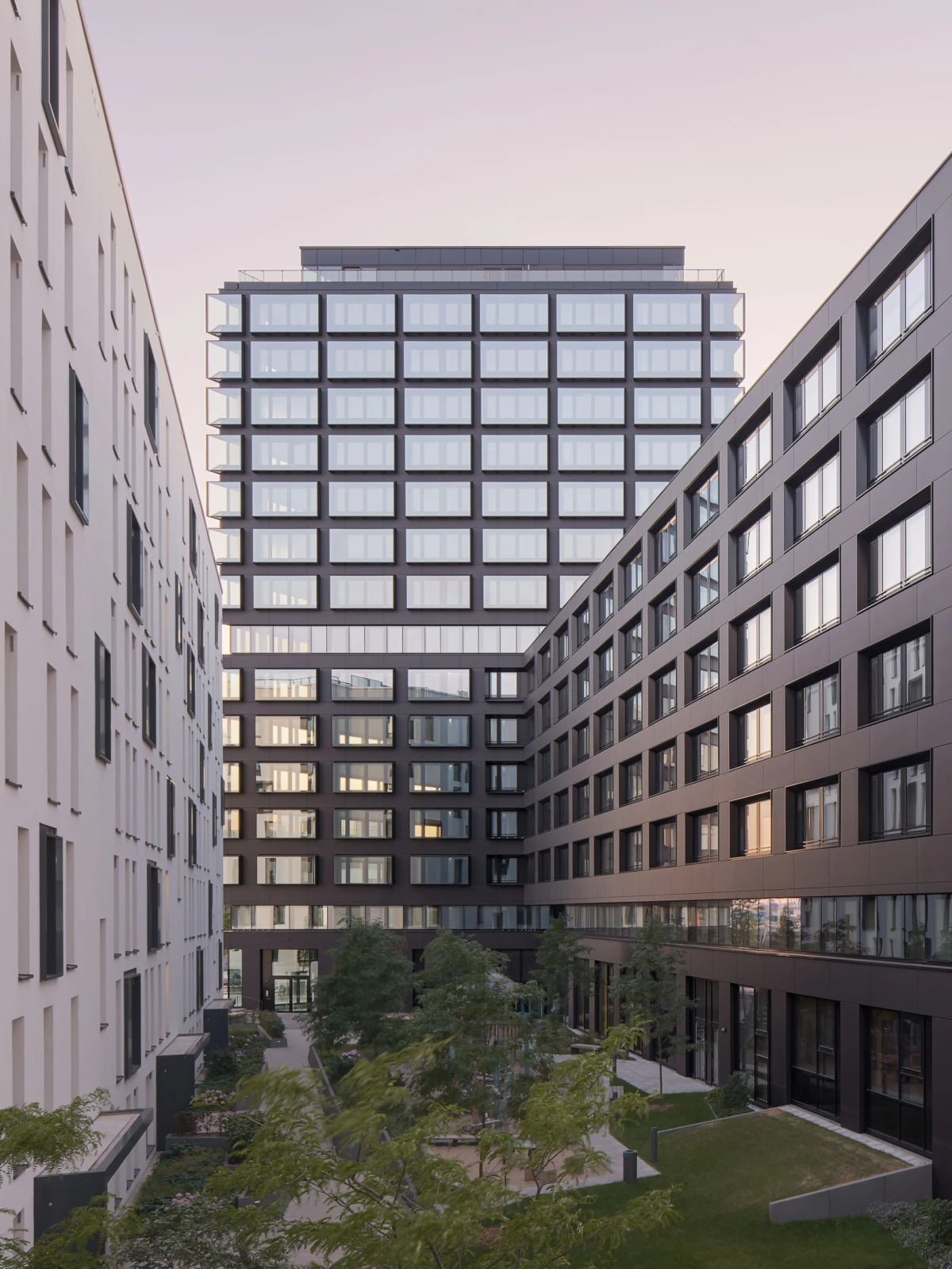 Courtyard between pale white and dark gray glass office blocks; trees, lawns, and winding paths under a pink sunset.