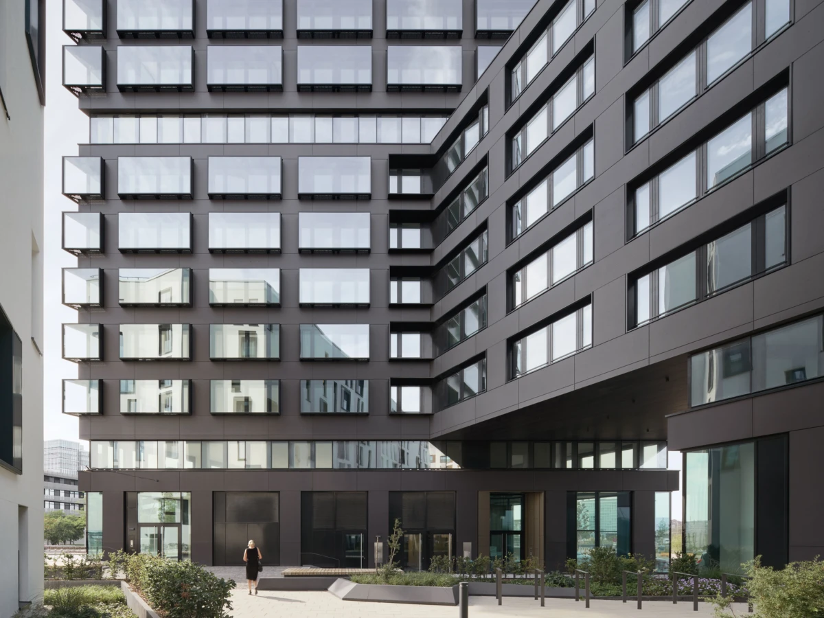 Person walking toward the entrance of a modern gray office building complex with glass windows in a courtyard.