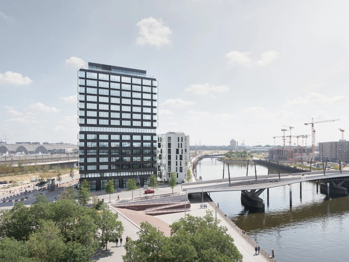Tall modern glass office building beside a river, with a bridge over the water, trees in foreground, and construction cranes in the distance.