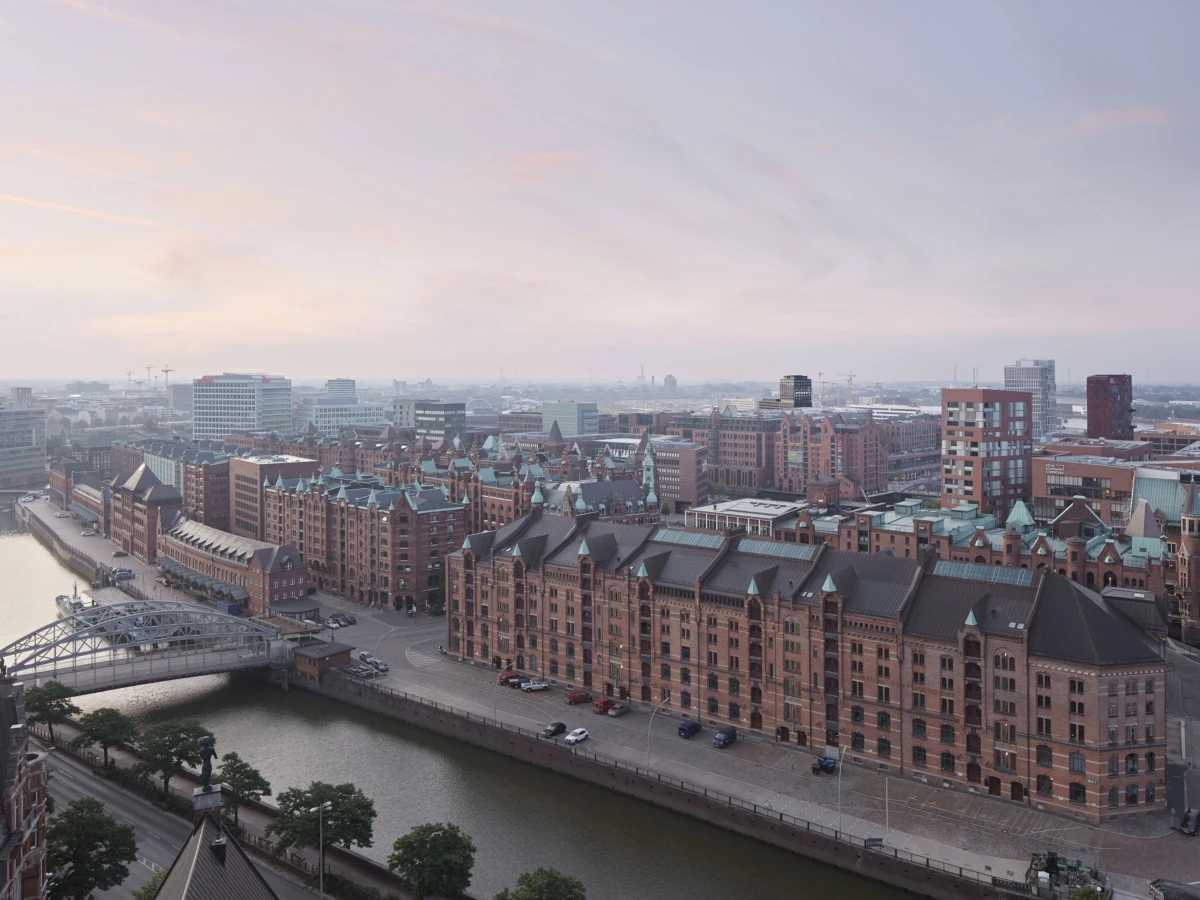 Aerial view of red brick warehouse district along a canal, with an arched metal bridge and riverfront road at sunset.