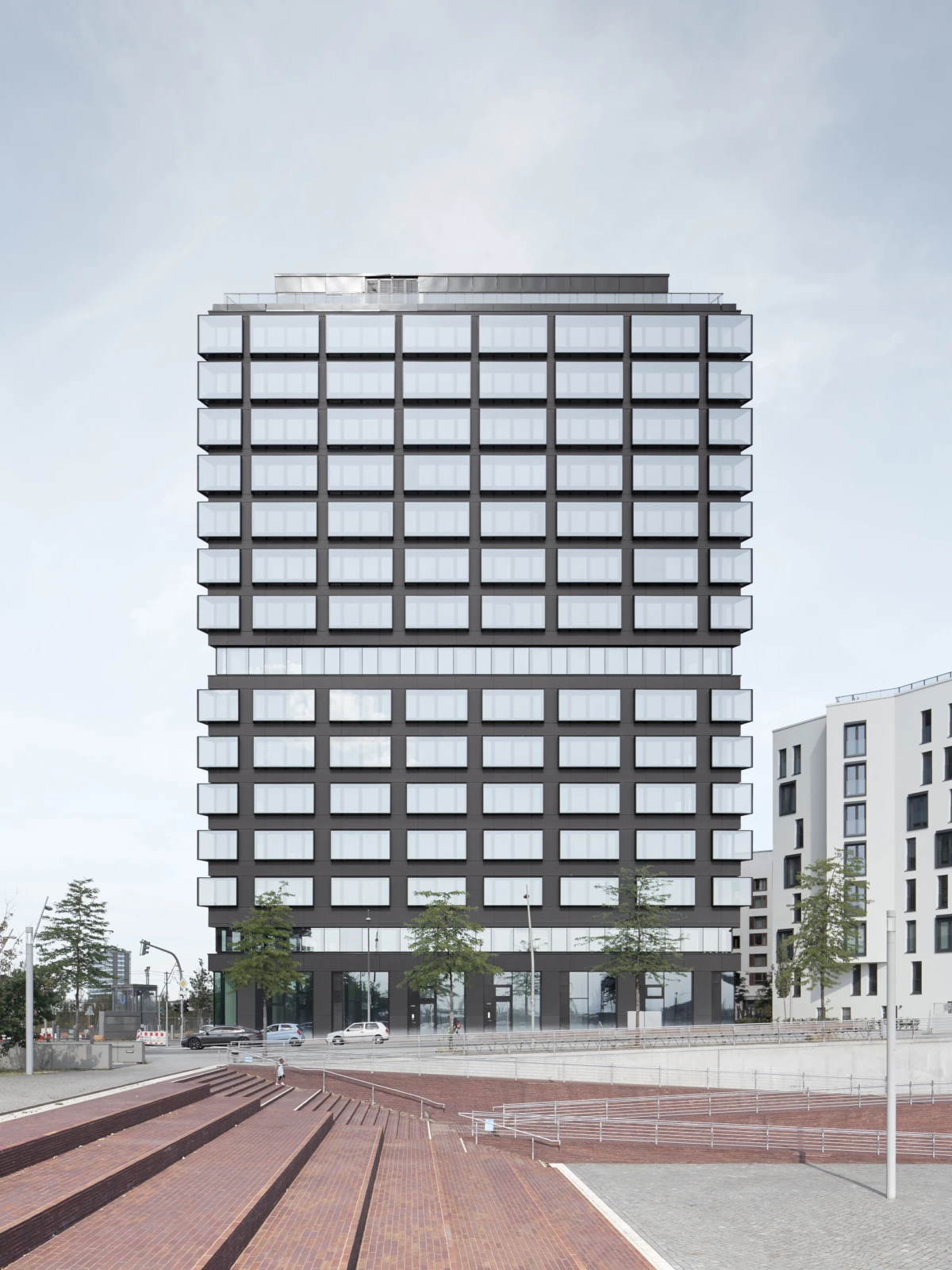 Tall modern office building with a glass facade in an urban plaza with brick steps and trees.